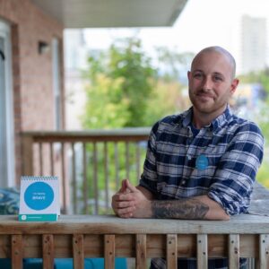 Greg Swaine smiling in a blue and white checked shirt, leaning against a wooden railing, hands folded in front of him with a small sign that says 